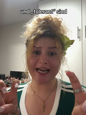 A woman with curly blonde hair and a green flower in it is looking directly at the camera with an excited expression, pointing her fingers together. Above her head, text is overlaid: "und „,tolerant“ sind". She is wearing a green shirt with white stripes and a gold necklace.