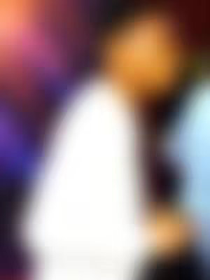 An out-of-focus image featuring a person in a white garment. The background has blurred colors of purple, orange, and blue, suggesting an indoor setting. The primary focus is a person, with a white surface dominating the left side of the image, where we have mostly seen the clothes or skin of the person.