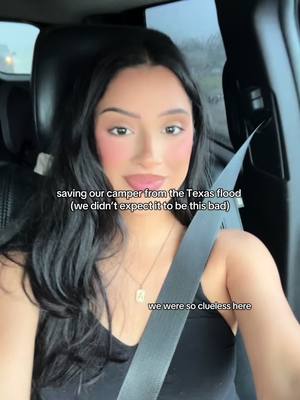 A woman with long black hair is seated inside a car, facing the camera. She has makeup on, including blush, and is wearing a black top and a necklace. A seatbelt is across her chest. Text overlays indicate the video's focus on a Texas flood and saving their camper.