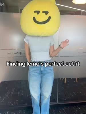 A person with a giant yellow lemon head costume is standing in front of a wall and wearing a white t-shirt and jeans. Text overlay at the bottom that says 'Finding lemo's perfect outfit'.