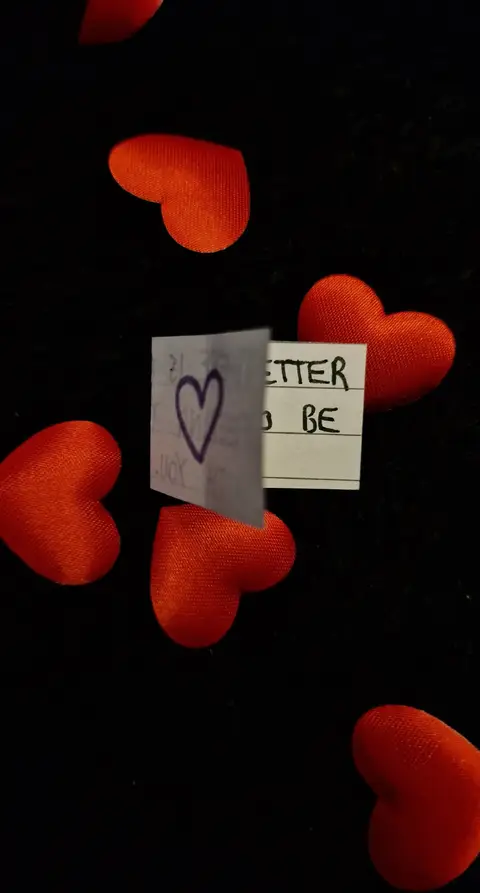 The image shows a composition of red fabric hearts against a dark background. In the center, a small, folded card is placed. It has a drawn heart and handwriting in black ink, spelling out the words 'I LOVE U' and 'it is BETTER to be' with blank lines below 'to be.' The arrangement suggests a romantic or sentimental theme, using the hearts as a visual cue for love.