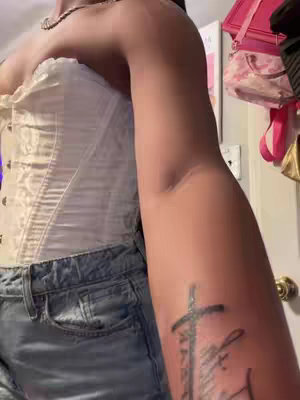 The image displays a person's torso and arm, with a white corset and denim shorts forming the upper body attire. The arm is tattooed with a cross and script. Details like a necklace and background elements such as a door are visible.