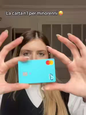 A person holding a turquoise credit card in front of their face with both hands. The background is a blurry interior setting with a door visible. The person is wearing a black top and has blonde hair.