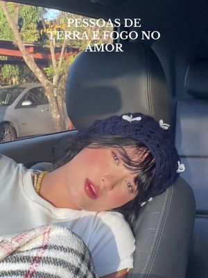 A young woman with fair skin and dark hair is seated in a car. She's wearing a white shirt and navy blue crocheted hat. Her cheeks are flushed, and she leans her head against the seat, conveying a thoughtful or slightly sorrowful expression. Text overlays the cover that reads: "PESSOAS DE TERRA E FOGO NO AMOR". The background shows a car interior.