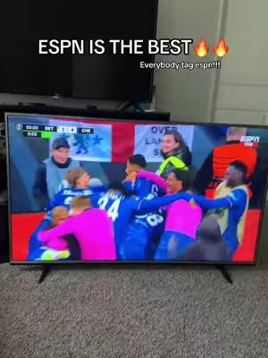 A television screen displays a soccer match with players in blue and pink uniforms celebrating a goal. Text overlays at the top promote ESPN. The background has a TV console and a grey carpet. The overall composition highlights the excitement of the sport.
