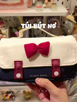 The image displays a close-up of a pencil case held by a hand. The pencil case is cream-colored on top with a navy blue base, and features a red bow in the center with two navy color tabs. The hand is visible, holding the lower part of the pencil case. The environment around appears to be objects on shelves or containers.