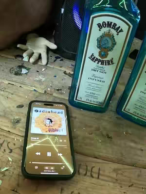 A close-up shot of a phone playing music, a bottle of Bombay Sapphire gin, and a white glove on a rough wooden surface. The phone displays a music app with the Radiohead song "Paranoid Android" playing. The gin bottle is teal with white labels, and a white glove lies nearby. The surface appears to be a wooden board.
