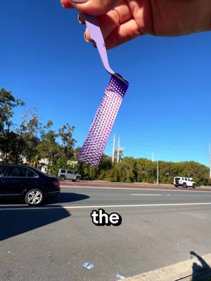 A clear, sunny shot with a hand holding a purple, textured strip against a vibrant blue sky. The scene includes a road with cars, greenery, and utility poles, suggesting an outdoor, urban environment. The composition is from a low angle, focusing on the strip in the hand with text at the bottom that says 'the'.