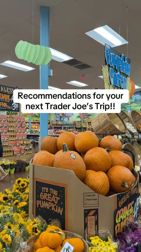 The cover photo presents a vibrant scene within a Trader Joe's store, focusing on a display of ripe pumpkins and related seasonal merchandise. A large box filled with multiple orange pumpkins dominates the foreground, with a 'Great Pumpkin' sign posted on the box while sunflower bouquets and colorful floral arrangements surround the pumpkin display. A green decorative element hangs from the ceiling, and the background hints at the store's aisles filled with products and signage. The image's composition is appealing and suggests seasonal grocery suggestions or themed food items.