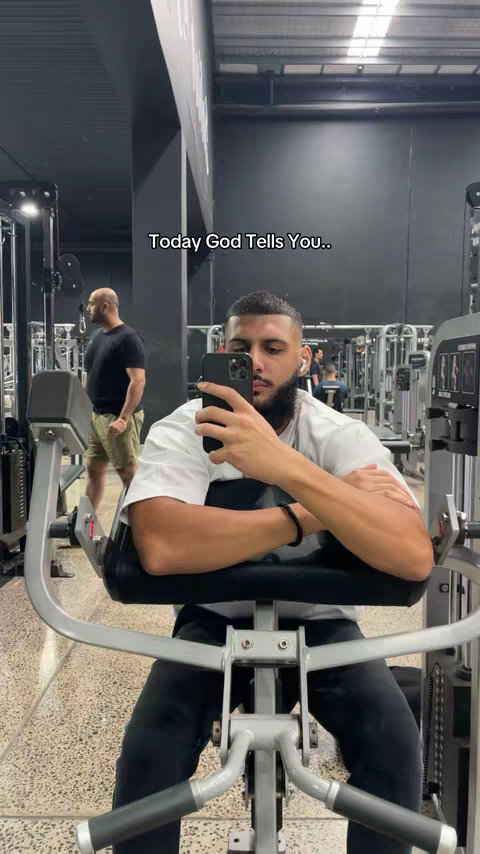 A man is shown taking a selfie in a gym. He is seated on a machine with his arms resting comfortably on the armrests. The background partially reveals other gym equipment and another person. The text overlay reads 'Today God Tells You....'