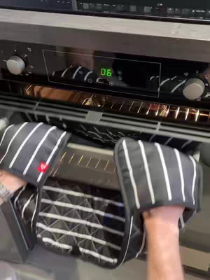 A first-person view shows a kitchen oven with the door open. Oven mitts with black and white stripes are being used to pull something out. The face of the oven is dark gray with a digital display showing the number '06' in green.