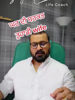 A man in a white shirt and glasses sits in a green chair against a blurred grey background. Text overlays in red and yellow Punjabi script.