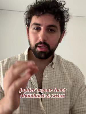 A man with dark curly hair and a beard is centered in the frame, speaking towards the camera. The background is a plain white wall. Overlaid text reads "Jupiter in your chart: Abundance & excess".