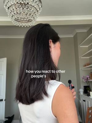 A woman with dark hair is seen from the back, facing away from the camera. She is wearing a white sleeveless top and holding a microphone over her shoulder. The background features a chandelier, a wall, and a glimpse of shelves and decor. Text overlayed on the image reads: "how you react to other people's".