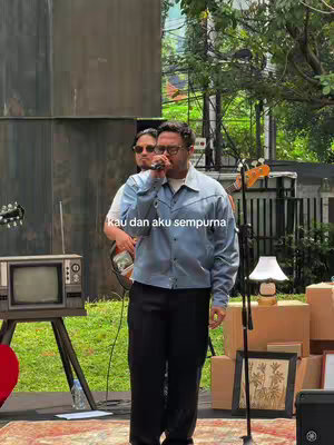 Two men are on a stage outdoors, in front of greenery and a stage-like structure. One man is singing into a microphone, and the other playing the guitar. The central figure wears a light blue jacket and black pants. Text overlay says "Kau dan aku sempurna".