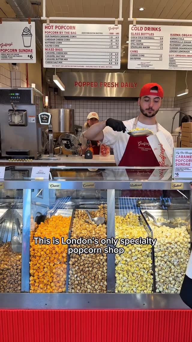 A video cover image featuring the interior of a popcorn shop with a male employee behind the counter serving popcorn. In the foreground, there are containers filled with various popcorn flavors like cheese, caramel, and salted. Above the counter, there are menu boards and signage for "Godfrey's" popcorn. The overlay text says, "This is London's only specialty popcorn shop".