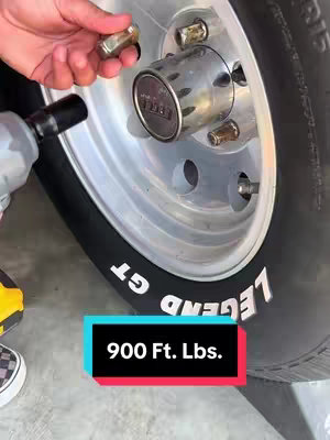 A close-up shot showing a tire and wheel with a mechanic's hand with a lug nut. In the foreground, an impact wrench. A graphic overlay shows the text "900 Ft. Lbs." over a blue and red shape.