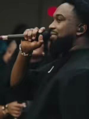A man with short, faded black hair and a trimmed beard is holding a microphone in his right hand. He's wearing a black outfit and an earbud, likely in a performance setting. The background is dimly lit and out of focus, with the man's face mostly in profile and focused on the microphone.