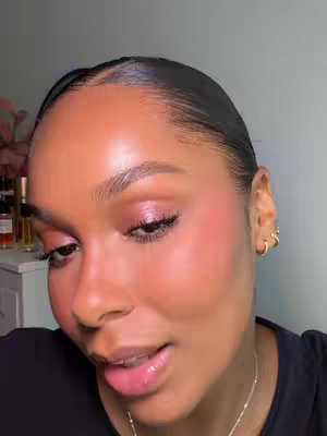 The cover image is a close-up of a woman's face, emphasizing her makeup. Her skin has a dewy, glowing appearance. She's looking slightly to the side, with her lips slightly open. Her hair is pulled back, styled in a sleek, glossy updo. She has gold hoop earrings and a delicate necklace. The background is out of focus. The overall colors are warm, with pink and peach tones dominating the makeup.