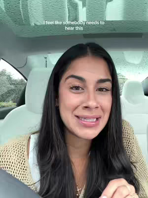 A woman with long, dark hair is seated inside a car, looking directly at the camera with a slight smile. She is wearing a light-colored top with a knit sweater and a delicate necklace. The background shows the interior of the car, including the headrests of the front seats and a glimpse of the overhead roof and windshield with rain drops.
