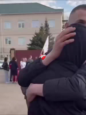 The image shows a moment of embrace between two people. The person on the left is partially visible with a black jacket, embracing another individual. The person being embraced seems to be holding something white. The background shows a building with a green roof, and indistinct figures. The lighting appears natural, creating a candid, emotional atmosphere.