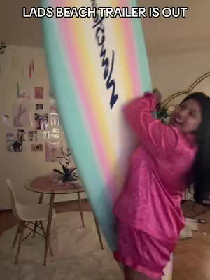 A woman in a pink robe holds a large, colorful surfboard against a wall. She smiles with excitement as she leans towards the board. In the background, there's a white chair, a round table, and photos on the wall, suggesting an indoor setting.