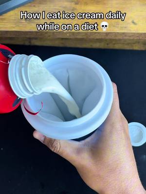 The image shows a top-down close-up of a person pouring a light-colored liquid from a red-topped bottle into a large white container. A dark surface is visible beneath the container. The hand holding the container is visible, and the focus is on the stream of liquid and the interior of the container.