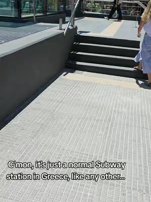 The image shows a descending view of a subway entrance in Greece. The entrance leads to dark stairs with a stainless steel handrail. The ground has a textured light-gray tiled pattern that contrasts against the darker shade of the surface around the stairs. A person in a white shirt and blue pants are on the right side. In the background, one person in walking by the entrance.