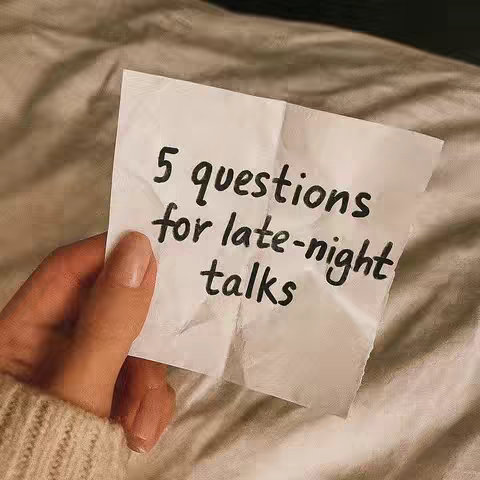 A close-up shot of a hand holding a crumpled piece of paper with handwritten black text against a soft, textured beige background, likely a bedsheet. The paper contains the words '5 questions for late-night talks'. The hand has painted nails and a neutral polish.