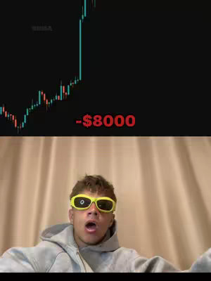The image shows a young man with yellow sunglasses in front of a neutral curtain. Behind him is a chart that seems to track the fluctuations of a trading stock. Below the chart, the text reads '-$8000', which is colored in bright red and presumably represents a financial loss. The man's expression is one of shock, with a wide mouth and open eyes, as if reacting to the value of the chart. The composition suggests a reaction video to a trading loss.