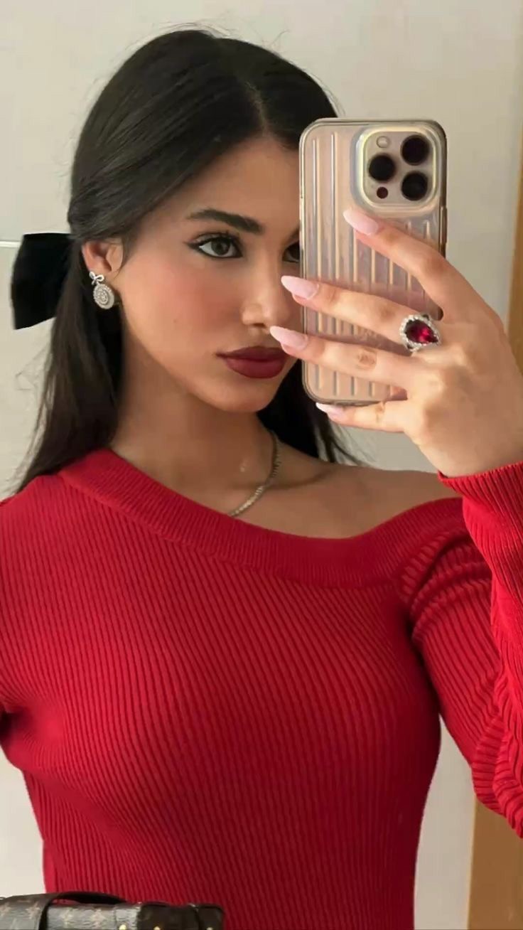 A selfie taken from a slightly high angle, featuring a young woman holding a phone up to her face. She has long dark hair styled back with a black bow. Her skin is fair, and her lips are painted with dark red lipstick. She is wearing a red off-the-shoulder sweater and a large, ornate ring with a red stone. The phone's screen partially reflects her face. Her nails are painted long and pink. The background is a cream-colored wall and a mirror.