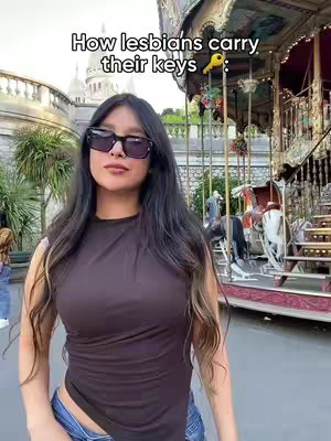 A woman with long, dark hair and sunglasses stands in front of a carousel. The background includes a blurred view of a building and people.