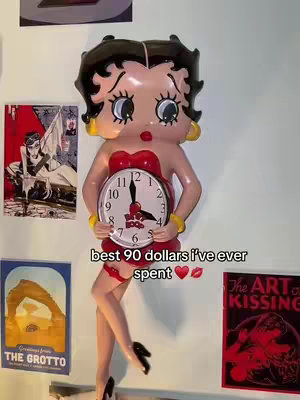A vertical cover image features a wall-mounted Betty Boop clock figure. The figure is in her iconic pose, holding a clock, and is set against a white wall adorned with other pictures and posters. Below the clock in the center of the image, the text "best 90 dollars I've ever spent" is displayed, with a heart and lips emoji to its side. The overall aesthetic is a blend of vintage and pop culture.