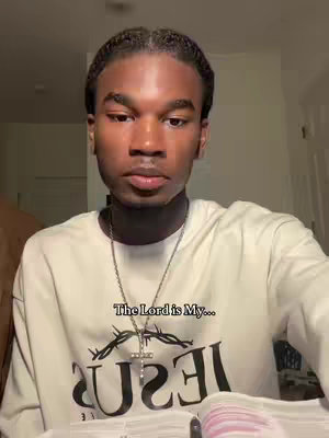 A young Black man is featured in a medium shot, positioned slightly off-center. He's wearing a white sweatshirt with the word "JESUS" visibly displayed in bold, dark letters across the chest, below a stylized crown of thorns. The man has short, neatly styled hair and is wearing a silver necklace with a cross pendant. He's holding pages of what appears to be a book or a bible. The background is softly lit, suggesting an indoor setting.