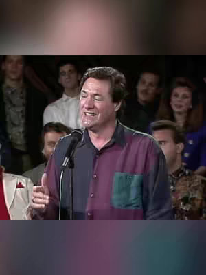 A man with dark hair and a patterned shirt speaks into a microphone, with a crowd in the background. The lighting is warm and the overall aesthetic is somewhat vintage.