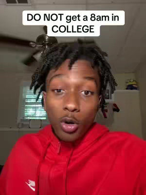 A young man with short black hair and a red hoodie is looking directly into the camera and speaking. Above his head is a white rectangular text overlay that reads, "DO NOT get a 8am in COLLEGE". The background appears to be a room with a ceiling fan and a window.