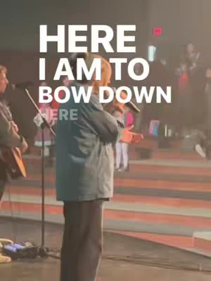 A person, likely a male, is standing on a stage, holding a microphone and singing or speaking. The background is softly lit, suggesting a performance or worship setting.