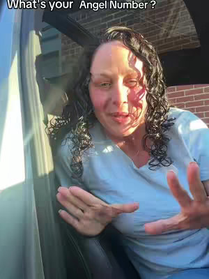 A woman with long curly hair, sitting in a car, gesturing with her hands. The background includes a brick wall and a lit window.