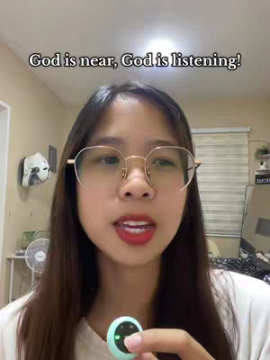 A person wearing eyeglasses is addressing the viewer in front of a beige wall. Above their head, white text states, “God is near, God is listening!” To their left, a decorative wall-mounted item and a fan are visible. The person holds a small, round object.