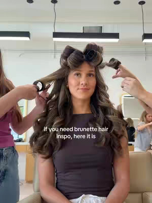 A woman with brown hair sits in a salon chair with rollers in her hair. Two stylists are attending to her hair. The image is well-lit, with a focus on the woman and her hair transformation. A text overlay provides more context.