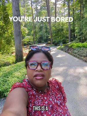A selfie taken in a park-like setting with a woman wearing glasses, casual attire, and text overlays. She has red-framed glasses and sunglasses up on her head. The background shows a path lined with trees, green bushes, and a well-lit pathway, with text overlays "YOU'RE JUST BORED" at the top and "THIS IS A" at the bottom, framing the subject's face. The overall composition suggests an informal, outdoor setting with a direct address to the viewer..
