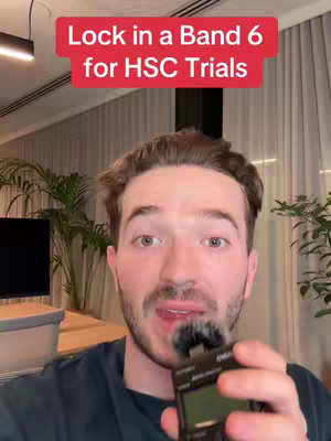 A man with a beard and short brown hair holds a small electronic device up to his mouth. He is slightly angled to the camera in a room with plants, a wooden table and a large window with blinds.