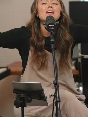 A woman with long, wavy brown hair is positioned in the center, singing into a microphone held in front of her. She is wearing a black long-sleeved shirt under a light tan jumpsuit. Her eyes are closed, and her mouth is open as if she is singing. A microphone stand and a tablet on a stand are in front of her. The background is blurred, suggesting a room with a desk or counter and a couple of monitors.