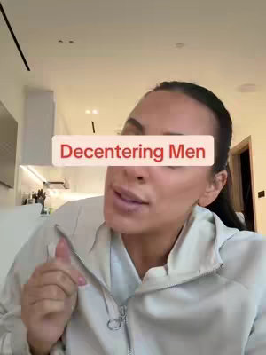 A woman in a light-colored jacket looks slightly to the side while speaking. A text overlay that says "Decentering Men" covers a portion of her face. The background is a blurred interior space with a white kitchen area.