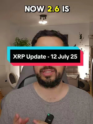 The image is a selfie-style video cover with a man centered in the frame. He is wearing a gray t-shirt. Above him, the text "NOW 2.6 IS" is displayed in a white, bold font. A prominent, colored rectangle with the text "XRP Update - 12 July 25" in white text sits over his mid-section. The man is against a blurred background, featuring interior elements like a light fixture, a plant, and what appears to be a door or wall decor.
