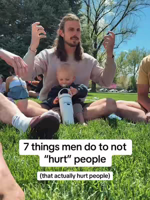 A man with long hair and a beard sits cross-legged on the grass, holding a baby on his lap. The man is looking towards the camera, raising his hands with open palms. Text in a white box at the bottom says, "7 things men do to not "hurt" people (that actually hurt people)". The background shows other people relaxing on the grass, trees, and a blue sky.
