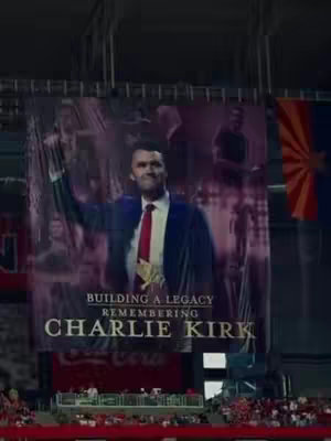 The image captures a banner at a memorial service. The banner showcases a large image of Charlie Kirk, a man in a blue suit and red tie, with the text "BUILDING A LEGACY REMEMBERING CHARLIE KIRK" below, in a bold, serif font. The background includes the venue's seating and flag.