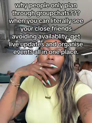 A selfie-style image with a woman in a yellow shirt as the central focus. The image is well-lit and appears to be taken in an office or public space, given the glimpse of a couch and other individuals in the background. She is holding her hand near her face with nails painted in a light green and white pattern, accessorized with multiple rings and bracelets, and a necklace. The background is blurred. The overall tone is casual and conversational but the subject’s direct gaze indicates that the content is directed straight at the viewer.