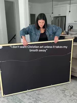 A woman is holding up a large framed canvas, displaying the back of the artwork. The canvas appears to be black, and held with both hands. Text is overlaid on the back of the canvas. She is wearing a denim jacket and is positioned in a home setting, with a kitchen visible in the background.