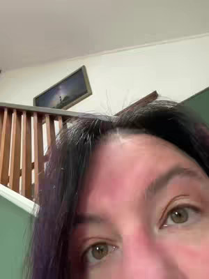 A face close-up of a woman with dark hair with a hint of purple, looking into the camera up close. The room's background is composed of white walls, a staircase with wooden railing, and a framed picture.